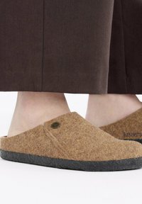 Pantofole in feltro marrone con suola in gomma nera, caratterizzate da una punta tonda, design slip-on e un piccolo logo rotondo sull lato.