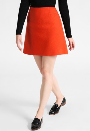 Woman wearing a bright orange A-line skirt, black long-sleeve top, and black patent leather loafers, standing with legs crossed.