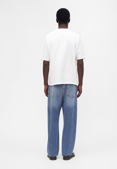 Homme debout de dos, portant une chemise blanche unie à manches courtes, un jean bleu ample et des chaussures noires sur un fond uni.