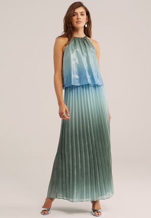Vrouw draagt een mouwloze, halternek, blauwgroene plissé maxi-jurk met een verloopkleur en zilverkleurige open-toe hakken, staand tegen een effen achtergrond.