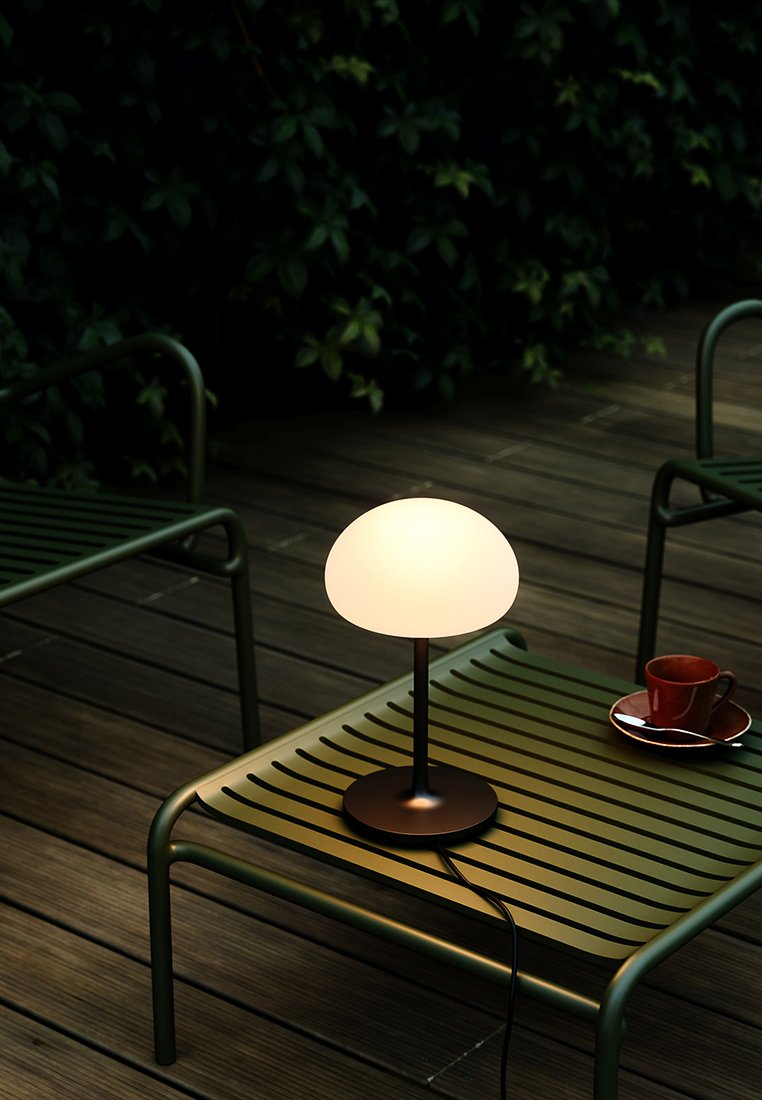 Tischlampe mit einem runden Opalglas-Schirm auf einem eleganten schwarzen Metallfuß, platziert auf einem grünen Latten Tisch neben einer roten Tasse und Untertasse.