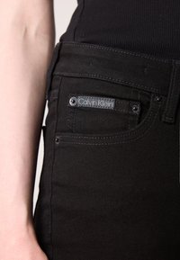 Blugi negri cu eticheta Calvin Klein pe buzunarul din față, purtați cu un top negru, arătând o parte din brațul și talia unei persoane.