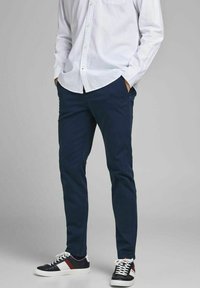 Pantaloni blu navy in cotone misto, design slim fit, indossati con una camicia button-up chiara e sneakers nere con dettagli rossi.