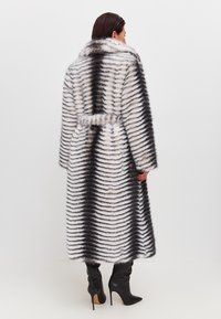 Lungo cappotto a righe realizzato in pelliccia sintetica, con motivi alternati bianchi e neri, colletto ampio e cintura annodata. Stivali neri con tacco visibili.