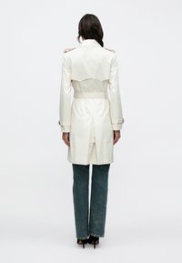 Fehér ballonkabát vállpántokkal, derékrészen övvel, és hátul sliccel, kék, bővülő szárú farmerral és fekete magassarkúval, hátulnézetben.