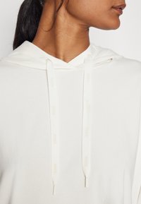 Närbild av en person som bär en vit hoodie med snoddar och en smidig tygtextur, vänd mot höger mot en neutral bakgrund.