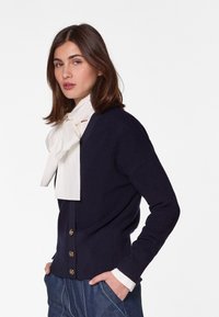 Marineblaues Cardigan mit Knopfverschluss, gefertigt aus strukturiertem Strickstoff, mit tiefem V-Ausschnitt und kontrastierendem weißen Fliegen. Kombiniert mit Jeanshosen.