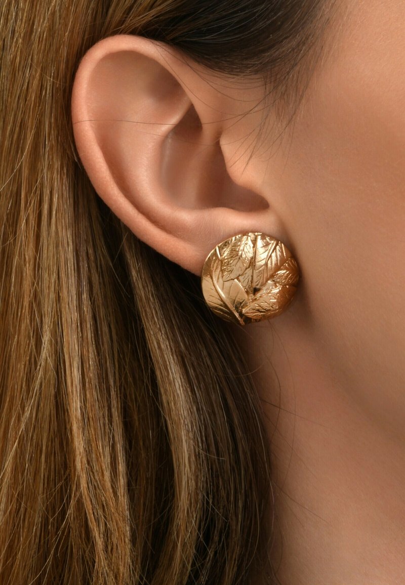Boucles d'oreilles rondes en ton or, avec une texture gravée de motifs de feuilles florales, posées sur un fond de couleur peau et des cheveux longs et raides.