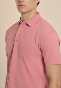 Camisa polo rosa de tejido texturizado, que cuenta con un cierre de dos botones, mangas cortas y un sutil logo bordado en el pecho.