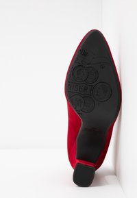 Røde ruskind højhælede sko med spids tå, der har en struktureret sort gummisål prydet med cirkulære mønstre og detaljer om mærkenavnet.