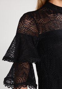 Sort blondebluse med høj hals, udslåede ærmer med bølgede kanter og detaljerede blomsterlige mønstre over hele stoffet.