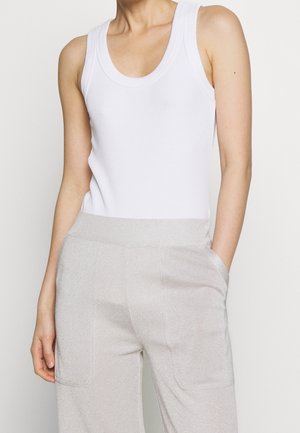 Kobieta ubrana w białą, bezrękawną prążkowaną koszulkę na ramiączkach oraz jasnoszare spodnie z wysokim stanem i widocznymi przednimi kieszeniami, z ręką w prawej kieszeni.