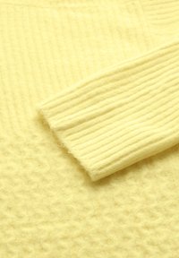 Gult stickat tyg med ribbad textur på ärmarna och en mjuk, fluffig yta, med ett bikakesmönster på kroppen.