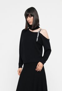 Black off-shoulder sweater in knit fabric with a ribbed hem. Features a strap with logo detailing. Model paired with a black skirt.