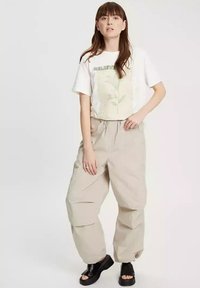 Biała, oversize'owa koszulka z zielonym napisem "BELIEVE" i graficznym wzorem kwiatowym, połączona z luźnymi beżowymi spodniami i czarnymi klapkami.