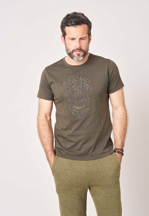 Man met baard die een olijfgroen t-shirt draagt met een bloemig schedelmotief en bijpassende broek, handen in de zakken, staand tegen een effen achtergrond.