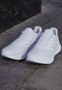Paar weiße Sportsneakers mit Schnürsenkeln, die auf einem grau gefliesten Boden in der Nähe einer dunklen Wand liegen.