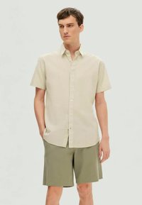 Világos bézs színű, rövid ujjú ing, gombos elejével, olíva zöld rövidnadrággal párosítva. Simább textúra, kényelmes vágás.