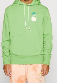 Zelená mikina vyrobená z bavlněné směsi, s přední kapsou, bílými šňůrkami a květinovým logem na hrudi. Nosená s světle oranžovými šortkami.