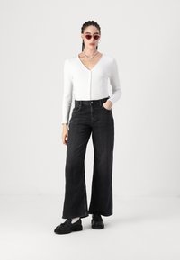 White ribbed long-sleeve top with a v-neck and button closure, paired with high-waisted black wide-leg jeans and chunky black shoes.