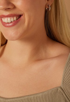 Nahaufnahme eines Frauenhalses und ihrer Schulter, die ein gestreiftes olivgrünes Gewebe zeigt; sie trägt kleine Creolen mit einer strukturierten Detailverarbeitung.