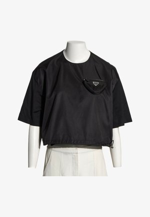 Chemise noire courte à manches courtes avec une petite poche zippée sur la poitrine gauche, portée sur un torse de mannequin blanc, associée à un pantalon blanc.
