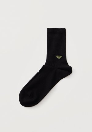 Chaussette noire côtelée à tige classique, ornée d’un petit logo d’aigle brodé en jaune, présentée sur un fond clair.