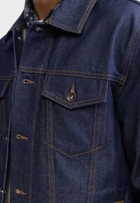 Chaqueta de mezclilla oscura con costuras naranjas brillantes, bolsillo en el pecho con un botón de metal y diseño de cuello clásico.