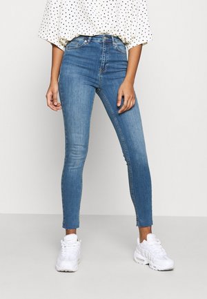 Kvinde iført højtaljede blå skinny jeans kombineret med en hvid top med polkaprikker og hvide sneakers, stående mod en ensfarvet baggrund.
