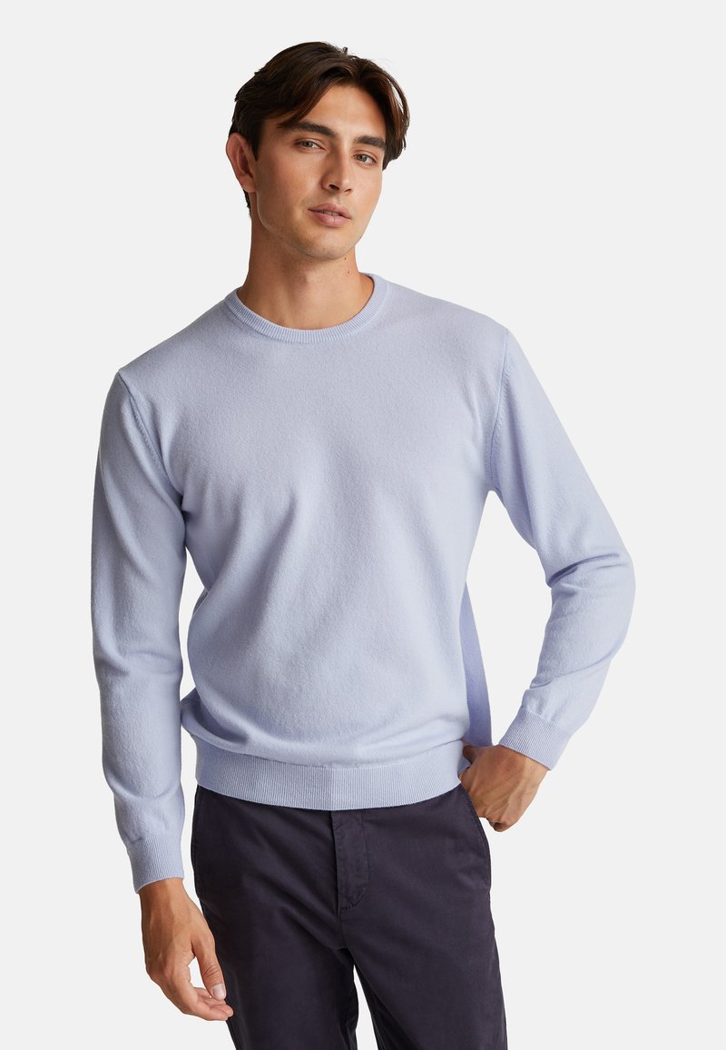 Mladý muž v světle modrém svetr s kulatým výstřihem a tmavých kalhotách, pózující s jednou rukou v kapse na jednobarevném pozadí.