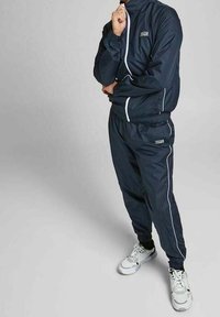 Chándal navy confeccionado en material ligero, con detalles en blanco, cierre de cremallera y puños elásticos. Combinado con zapatillas deportivas blancas.