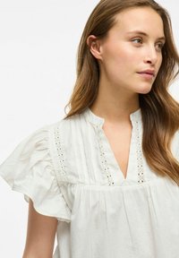 Blusa bianca leggera con scollo a V, maniche corte a volant, dettagli in pizzo, pieghe verticali e vestibilità ampia.