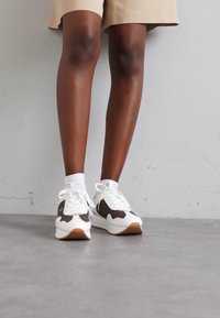White sneakers with brown accents, featuring a textured upper, round toe, and thick sole. Laces are tied at the front, complemented by white socks.