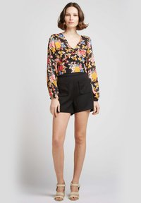 Blouse à manches longues florale avec une base noire, ornée de fleurs colorées. Associée à un short noir, le modèle porte des sandales à talons nudes.