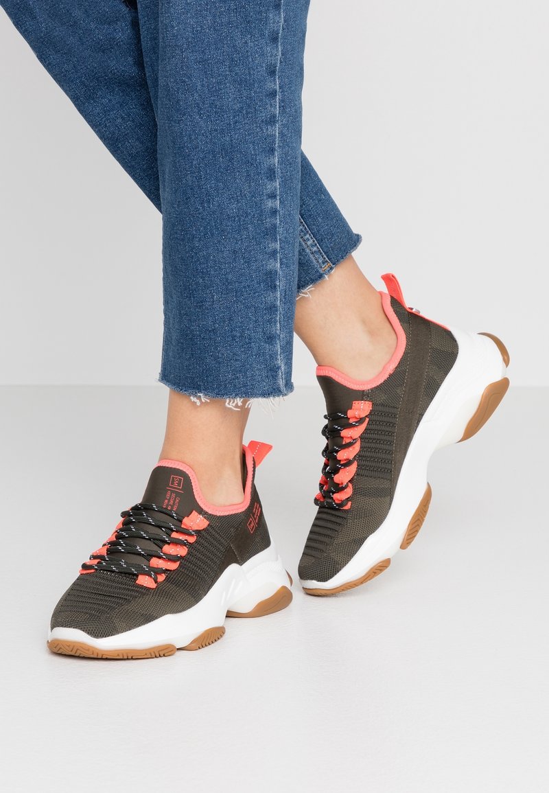 Tummanoliivinvihreät tennarit oransseilla yksityiskohdilla, joissa on neulottu päällinen, mustat nauhat ja valkoinen välipohja kumisella ulkopohjalla.