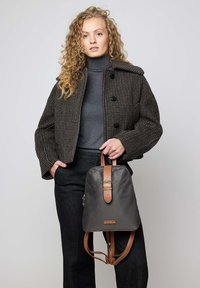 Zaino in nylon grigio con cinturini e fibbia in pelle marrone, caratterizzato da una texture liscia e un design minimalista, indossato da una modella con cappotto a quadri.