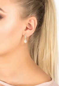 Arany tónusú, csepp formájú fülbeval ók, rózsaszín kövekkel és átlátszó díszítéssel, sima, fényes felülettel.