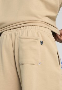 Beige joggerbukser lavet af blødt stof, med elastisk talje, sidelomme og blå detaljer nær lomme kanten.