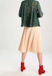 Blouse verte transparente à motif floral, jupe plissée crème, et bottines rouges à la cheville. La blouse possède un col rond et des manches longues.
