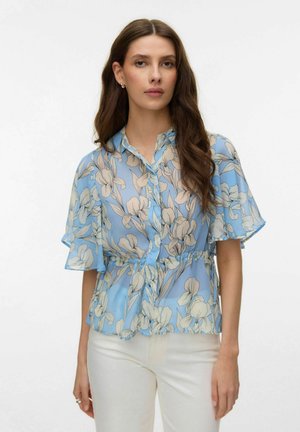 Femme portant un chemisier bleu à motifs floraux avec des manches volantées et un pantalon blanc, debout devant un fond blanc uni.