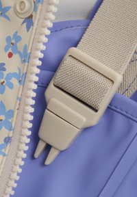 Verstelbare beige band met een plastic gesp, bevestigd aan een stof met een bloemenpatroon in crème en lichtblauw, tegen een paarse achtergrond.