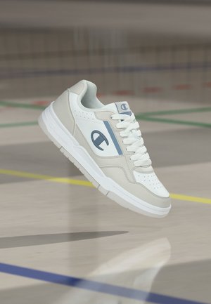 Valge ja hall sneaker sinise logoga hõljumas sise-spordiväljaku põranda kohal, kus on värvilised jooned.