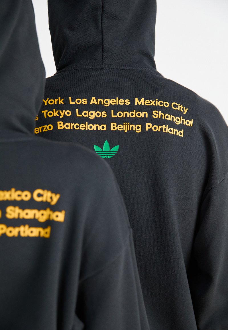 Czarna bluza z kapturem z żółtymi nazwami miast nadrukowanymi z tyłu oraz zielonym logo Adidas. Miękki materiał i luźny krój.