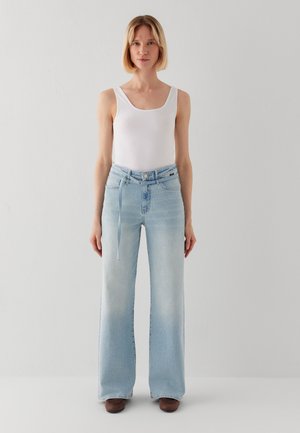 Frau steht vor einer weißen Wand, trägt ein weißes ärmelloses Oberteil, hellblaue hoch taillierte weite Jeans und braune Schuhe.