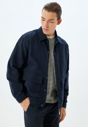 Chaqueta azul marino hecha de un tejido resistente, con cuello de punta, cierre frontal con botones y dos bolsillos laterales. Usada sobre una camisa a rayas.