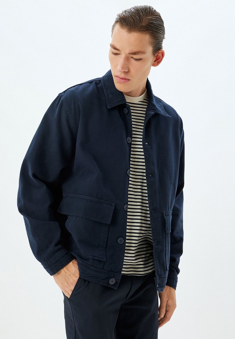 Giacca blu navy realizzata in tessuto resistente, dotata di colletto a punte, chiusura frontale con bottoni e due tasche laterali. Indossata sopra una camicia a righe.