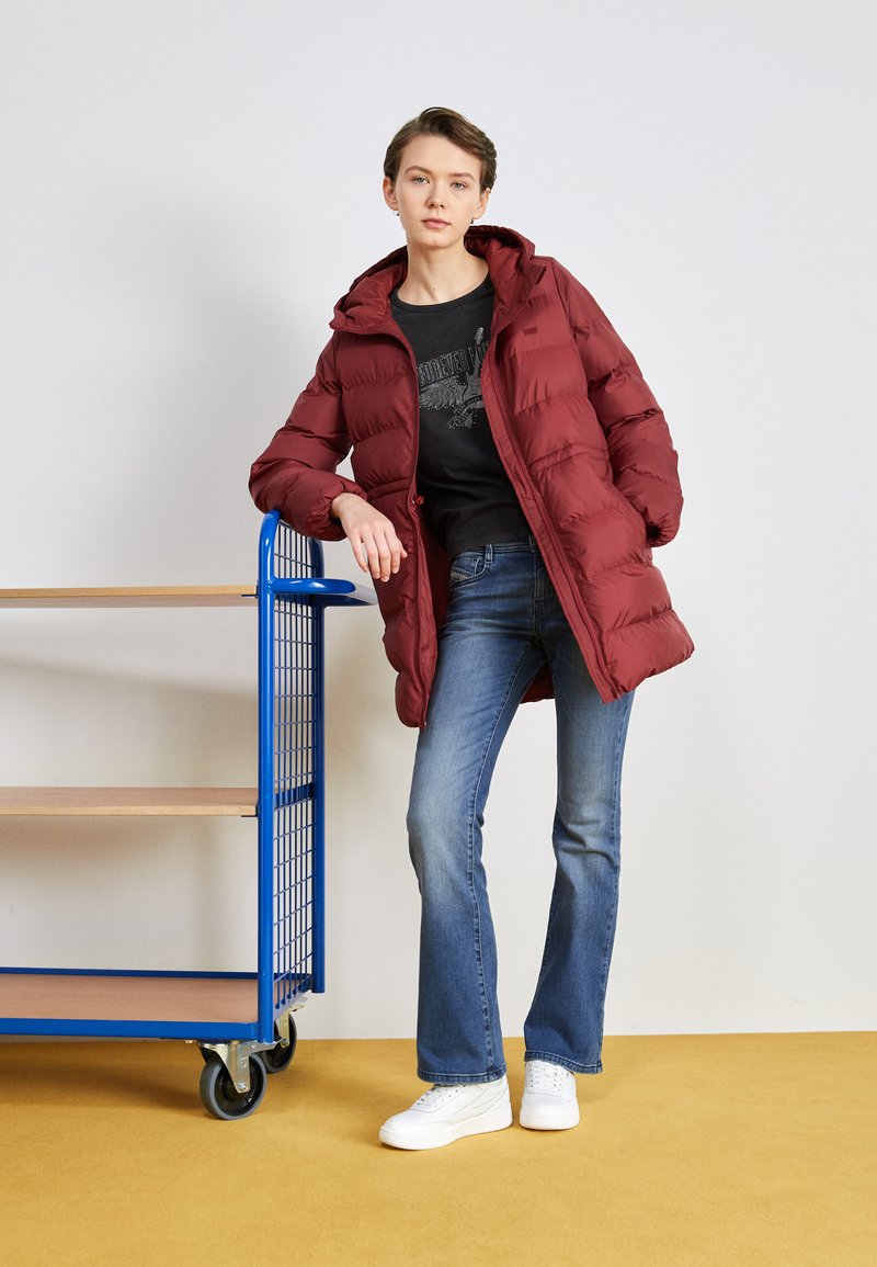 Cappotto imbottito bordeaux con cappuccio, caratterizzato da un design trapuntato e testurizzato. Indossato sopra una maglietta nera con grafica, abbinato a jeans svasati e sneakers bianche.