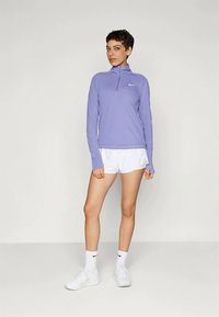 Ανοιχτό μωβ μπλούζα με μακριά μανίκια, μισό φερμουάρ στον γιακά και λογότυπο, συνδυασμένη με λευκά σορτς. Το μοντέλο φορά λευκά παπούτσια και κάλτσες.