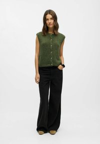 Pull sans manches en tricot vert avec des boutons sur le devant, associé à un pantalon large noir et des chaussures slip-on marron. Design simple et texturé.