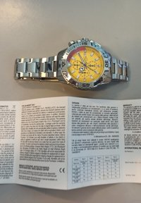 Orologio in acciaio inossidabile argento con quadrante giallo, caratteristiche cronografiche e bracciale a maglie; include il manuale di garanzia nelle vicinanze.